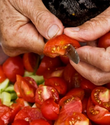 Old grandmother is preparing salad closeup. Old wrinkled hands of grandmother chop vegetables on summer salad. Grandmother prepares food for grandchildren close-up. Grandma cuts tomatoes